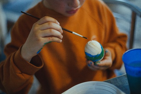 child decorates an Easter egg in the colors of the rainbow. Boy holds an egg and paints it with brush. Preparing for celebration of Easter.の写真素材