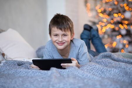 Happy handsome boy with a tablet in hands. child plays computer games on tablet. boy lies on bed opposite Christmas tree before Christmas. Black Friday and Cyber Monday Online Shoppingの写真素材