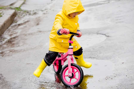 little girl in yellow raincoat rides on bike ride through puddles in rain. Rainy weather in spring. child learns to manage two-wheeled transport. Learning to ride on balance bikeの写真素材