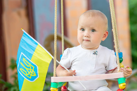 baby with Ukrainian flag sits on swing in park on walk. Child celebrates Ukraine's independence day at homeの写真素材