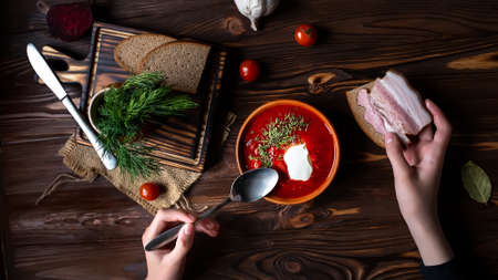 Hands holding a spoon and piece of bacon sandwich. Traditional Ukrainian borsht, red vegetable and meat soup or borscht with smetana on wooden background. Slavic dish with cabbage, beets, tomatoesの写真素材