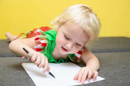girl writes a letter to Santa Claus. A child in Elf costume lies against yellow wall and makes wishes for Christmas. New Year 2022 concept.の写真素材