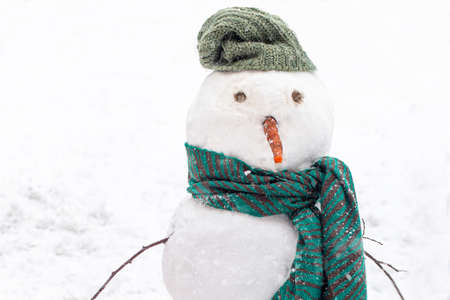 Snowman on background of snow drifts during snowfall in winter. snow figure with green knitted scarf and hat.の写真素材