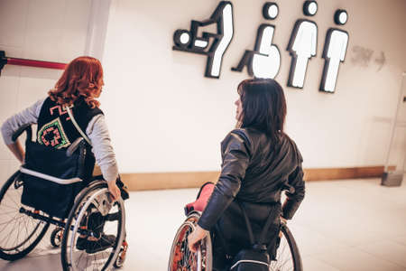 Two girls in wheelchairs near the entrance to the public toilet in the shopping center equipped for people with special needs. Toilets for special needs Person.の写真素材