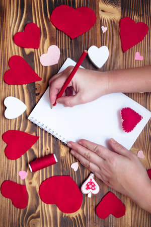 Hands writing down Valentines Day greetings in a blank notebook. Felt red hearts on a wooden tableの写真素材