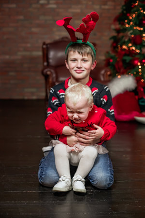 The brother holds his sobbing sister in his arms. New Year's family photo shoot. Brother and sister waiting for Christmas near the tree with lights in red clothesの写真素材