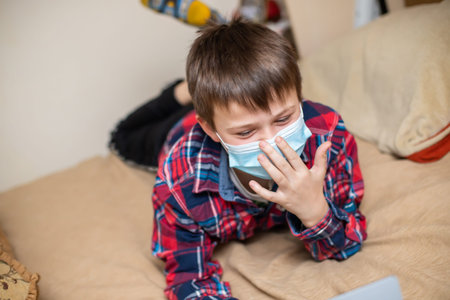 teenage boy in protective medical mask coughs in fist. child remotely does lessons lying on bed near laptop. problem of modern children. Kid and screens.の写真素材