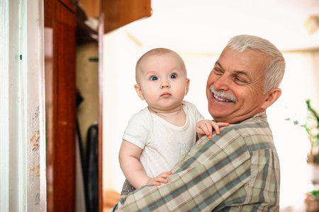 A happy smiling grandfather plays with his little grandson in a room against the window. An old man and a child up to a year oldの写真素材