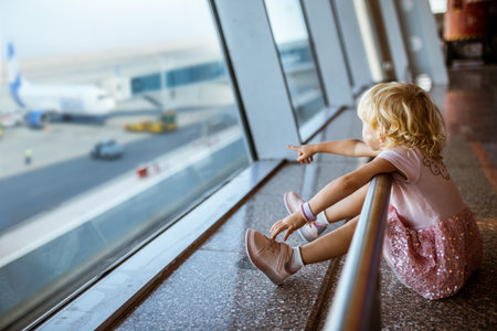 little girl sits by window at airport and points finger at plane. child in airport departure lounge. Girl sitting on floor waiting to board the planeの写真素材