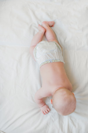 The baby lies on her stomach on a white sheet on the bed view from above. Newborn baby in diaper rests before bedtimeの写真素材