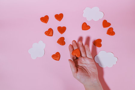A hand holding one red heart. Red hearts among paper clouds on a pink background. Conceptual background for Valentine's Day or Women's Dayの写真素材