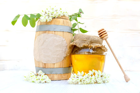 Fresh liquid acacia honey harvest 2023 in a wooden barrel and a transparent jar with a canvas napkin on the lid. Branches of flowering white acaciaの写真素材