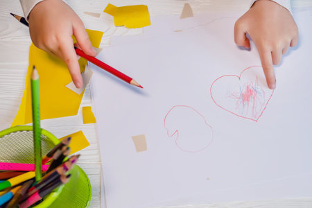 The hands of a small child point to a heart drawn by a child on white paper. top view. Mother's Day...