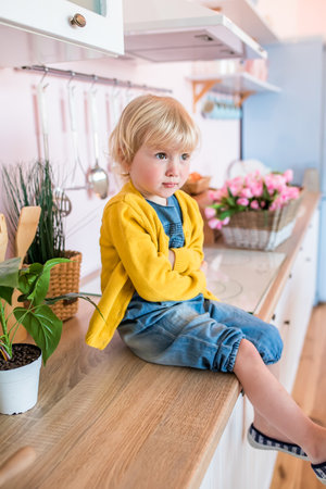 A little serious unhappy girl sits on the kitchen table with folded arms in front of her. An offended child in a yellow jacket.の写真素材
