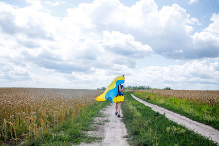 flag of Ukraine is on the shoulders of a little Ukrainian girl. Happy Ukrainian child in free Ukraine without war runs along a rural road in a field of wheat field.. International Mother Language Day.の写真素材