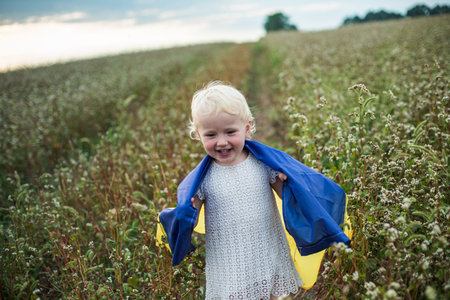 Proud Ukrainian little girl stands on field of mature buckwheat in a yellow-blue flag. day of Ukraines insability. Selective focus on flag.の写真素材