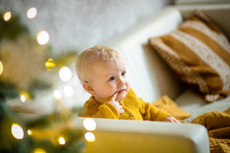 Little girl with finger in mouth in front of fur-tree with lights and gold decor. surprised girl....
