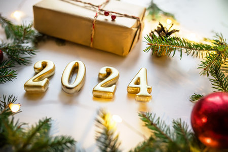 Golden numerals 2024 on white background with craft gift, christmas tree branches and red decorations and christmas lights.の写真素材