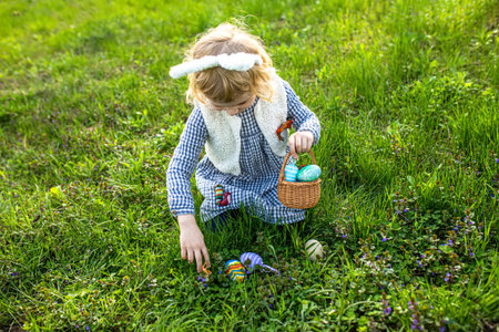 young girl, adorned in bunny ears, celebrating Easter by playfully collecting painted eggs in a...