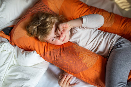 A Small girl is lying on top of a bed, with an orange pillow underneath her head. She appears relaxed and comfortable, with her eyes closed. The bed is neatly made with a white bedsheet.の写真素材