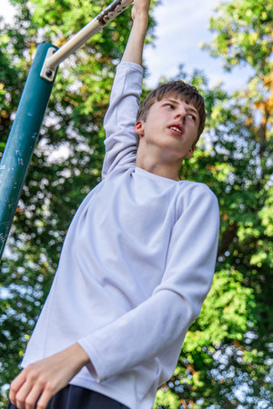 A young boy in a white long-sleeved shirt is exercising on a pull-up bar outdoors. He is pulling himself up, looking towards the sky. The background is a blurry green of trees.の写真素材