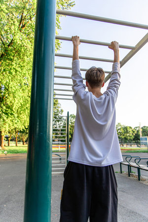 A young man in a white shirt and black shorts is hanging from monkey bars in a park. He is using the...
