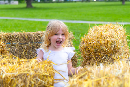 A young girl with blonde hair is having fun playing in a pile of hay bales. She is wearing a white shirt and is smiling brightly at the camera. The hay bales are surrounded by lush green grass, and the sky is clear and blue.の写真素材