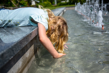 A young girl with long, blonde hair leans over the edge of a fountain, reaching her hand into the water. The water is sparkling and clear, and the girls expression is one of curiosity and delight. The fountain is surrounded by greenery and the sunlight is shining brightly.の写真素材
