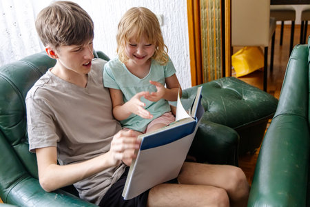 A teenage boy, sitting in a green leather armchair, reads a book to his younger sister who is sitting in his lap. The girl is pointing at the book and seems engaged in the story.の写真素材