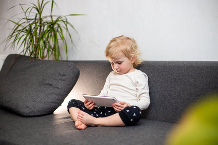 A young girl with blonde hair sits on a grey couch, legs crossed, and uses a white tablet. She is wearing a white long-sleeved shirt and black pants with white triangles. A green houseplant is visible to the right, slightly out of focus.の写真素材