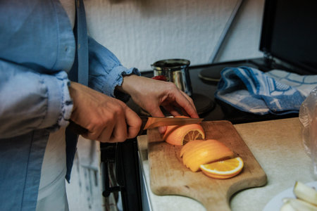 A person is skillfully slicing fresh citrus fruits, including oranges, on a wooden board. The kitchen has a warm atmosphere, filled with morning light.の写真素材
