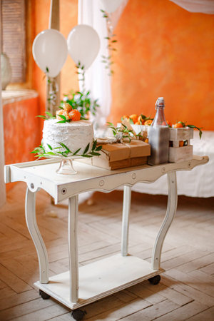 A white serving cart with a white cake topped with oranges and greenery sits on a white cake stand. The cart is also decorated with boxes, a glass bottle, and more oranges. The cart is on a wooden floor, and there is a white bed and orange wall in the background.の写真素材