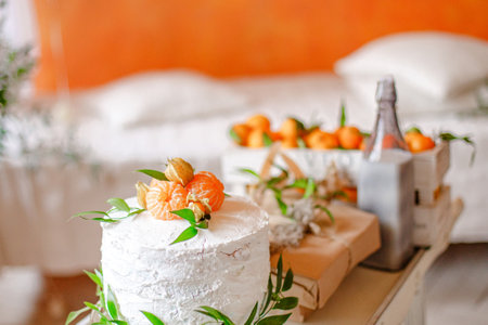 A white frosted cake adorned with mandarin orange slices and greenery sits on a table. The cake is in the foreground with a blurred background showing a white bedspread, pillows, and a box of oranges.の写真素材