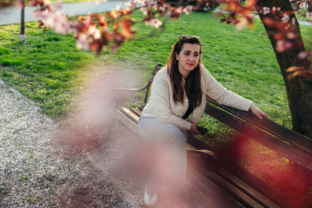A young woman relaxes on a wooden bench in a park, enjoying the warm sunshine and vibrant blossoms of spring. The atmosphere is peaceful and lively, perfect for unwinding.の写真素材