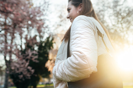 A woman walks through a park at sunset, enjoying the beauty of blooming trees and the warm golden light. The atmosphere is calm and serene, capturing a peaceful moment.の写真素材