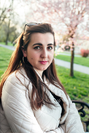 A young woman enjoys a moment of relaxation on a bench in a park adorned with blooming trees. The setting sun casts a warm glow over her serene expression, enhancing the beauty of spring.の写真素材