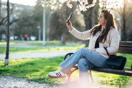 Woman taking a selfie on a park bench surrounded by blooming trees in early spring sunlightの写真素材