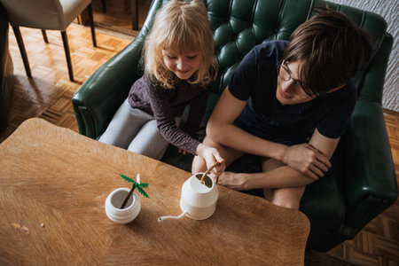 Little girl and teenage boy having fun indoors while watering a small artificial plant, enjoying quality sibling time.の写真素材