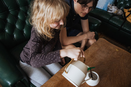 A child is engaged in imaginative play, pouring water from a teapot into a small plant pot while sitting at a wooden table. An adult observes nearby in a comfortable living room setting.の写真素材
