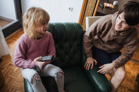 Two siblings lounge in a cozy living room. One child holds a remote while sitting on a green couch, and the other engages in conversation, creating a relaxed atmosphere.の写真素材
