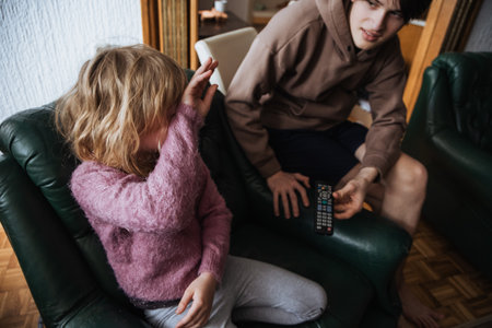 A young girl sits on a green chair, covering her face, while her older brother, in casual attire, sits nearby with a remote control in hand. They are in a comfortable living room.の写真素材
