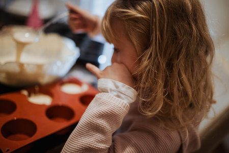 A young girl sits in the kitchen, focused on pouring batter into a silicone mold. Her expression shows excitement as she shares this baking experience with family.の写真素材