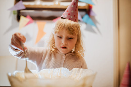 A young child in a party hat stirs cake batter in a bowl during a family celebration. Colorful decorations and a festive atmosphere create a cheerful mood for the special occasion.の写真素材