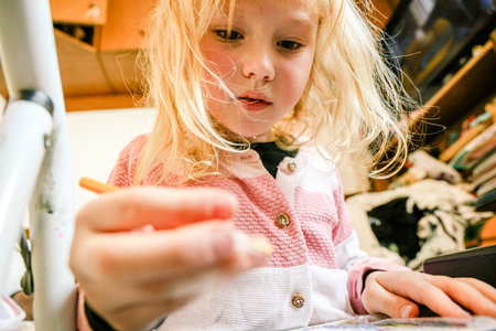 Blonde girl focused on creating diamond mosaic artwork at home. Concentrated expression, crafting with small gems and tools on a black table in a cozy room.の写真素材