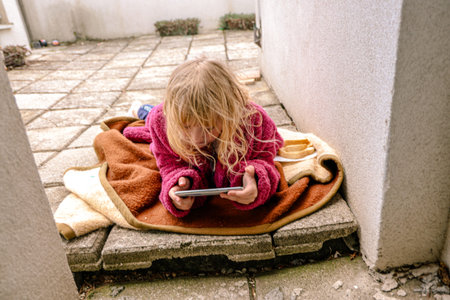 A young child with messy hair is lying on a blanket outside, focused on a smartphone. The setting is a residential calm area with clean paved stones and minimal distractions.の写真素材