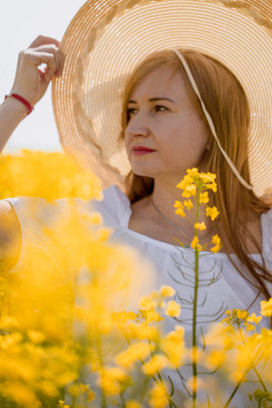 A woman in a white dress and straw hat walks peacefully through a rapeseed field, enjoying the beauty of nature on a sunny day.の写真素材
