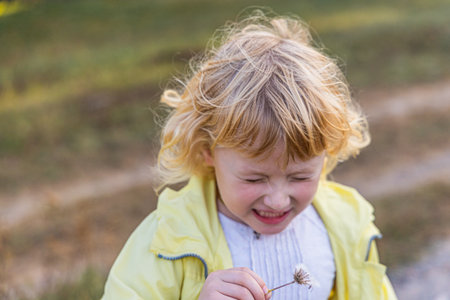 A cheerful child with curly blonde hair smiles while holding a small flower, surrounded by a lush grassy landscape under soft afternoon sunlight, capturing a moment of pure joy.の写真素材