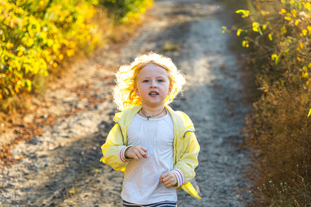 A young child in a light jacket dashes along a dirt trail surrounded by golden grass and green foliage. The scene captures the carefree spirit of childhood in a serene outdoor setting.の写真素材