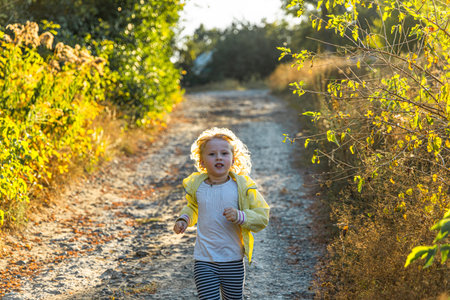 A young child in a light jacket dashes along a dirt trail surrounded by golden grass and green foliage. The scene captures the carefree spirit of childhood in a serene outdoor setting.の写真素材