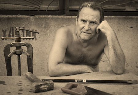 An elderly man thinking sitting on the bench at the table.On the table are locksmith toolsの写真素材